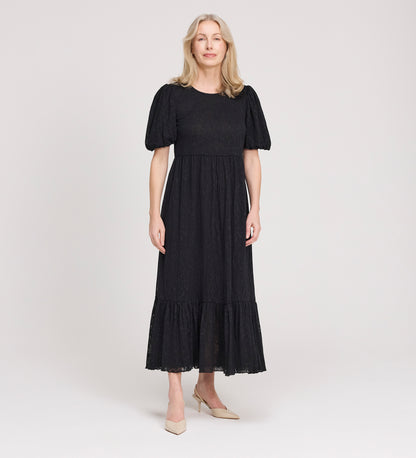 A woman with light hair stands against a plain background, wearing the Yasmine Black Jersey Midi Dress with puff sleeves and a ruffled hem, paired with beige pointed-toe heels.