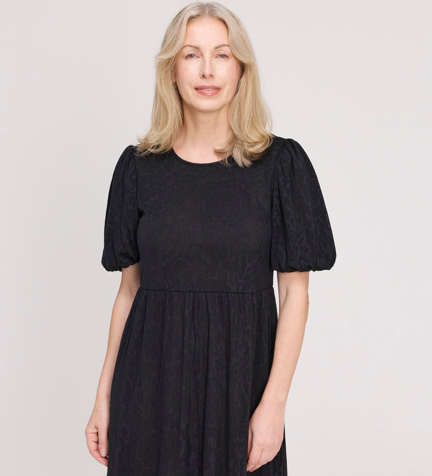 A woman with light blonde hair smiles gently at the camera, wearing the Yasmine Black Jersey Midi Dress against a plain light background.