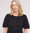 A woman with light blonde hair smiles softly at the camera while wearing the Yasmine Black Jersey Midi Dress, standing against a plain light background.
