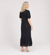 A woman with blonde hair faces away, wearing the Yasmine Black Jersey Midi Dress featuring puff sleeves and a ruffled hem, paired with beige heeled shoes against a plain light background.