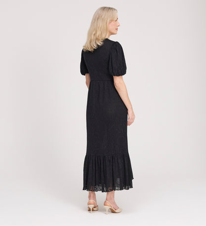 A woman with blonde hair faces away, wearing the Yasmine Black Jersey Midi Dress featuring puff sleeves and a ruffled hem, paired with beige heeled shoes against a plain light background.