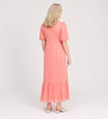 A woman with light hair faces away, wearing the Yasmine Coral Jersey Midi Dress—relaxed fit, ruffled hem, short sleeves—paired with white flats on a plain light background.
