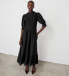 A woman wearing the Amelia Black Chiffon Dress with puff sleeves stands against a plain white wall, one hand on her hip. She pairs the textured midi dress with pointed burgundy heels and has her hair pulled back.