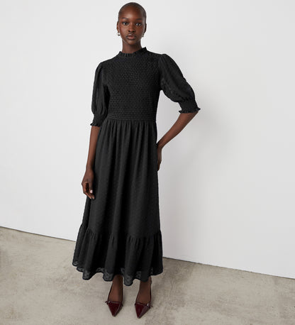 A woman wearing the Amelia Black Chiffon Dress with puff sleeves stands against a plain white wall, one hand on her hip. She pairs the textured midi dress with pointed burgundy heels and has her hair pulled back.