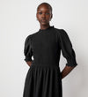 A woman stands confidently against a white background, wearing the Amelia Black Chiffon Dress featuring a textured fabric, puff sleeves, and a high neckline.