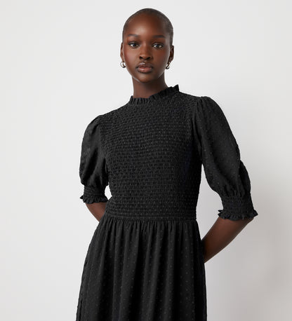 A woman stands confidently against a white background, wearing the Amelia Black Chiffon Dress featuring a textured fabric, puff sleeves, and a high neckline.