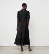 A person with closely cropped hair stands facing a white wall, wearing the Amelia Black Chiffon Dress—an ankle-length piece with puff sleeves and ruffle details—paired with sandals.