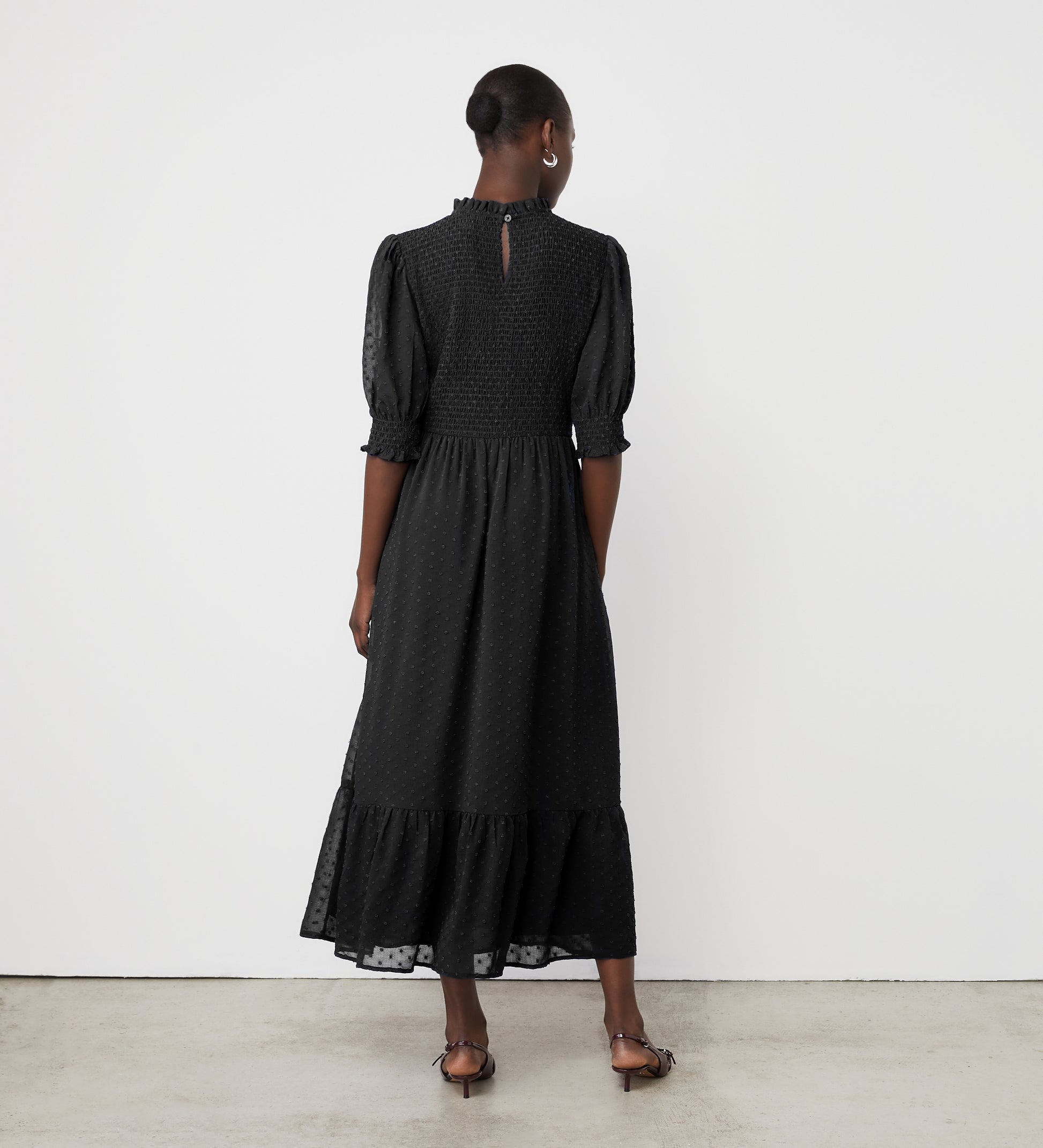 A person with closely cropped hair stands facing a white wall, wearing the Amelia Black Chiffon Dress—an ankle-length piece with puff sleeves and ruffle details—paired with sandals.