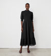 A woman stands against a white wall wearing the Amelia Black Chiffon Dress, featuring puff sleeves and a ruffled hem. She completes her look with pointed dark heels and exudes a confident, composed expression.
