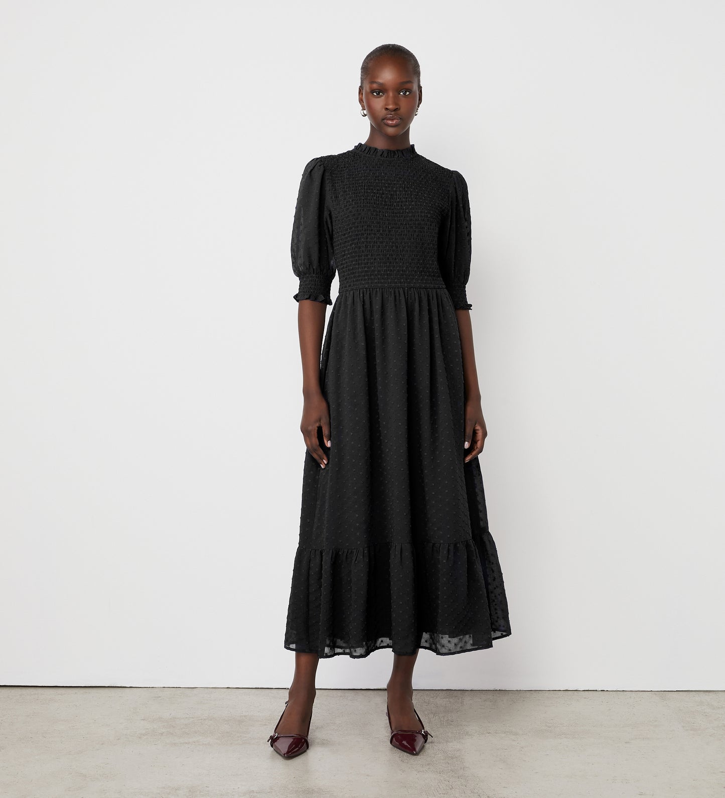 A woman stands against a white wall wearing the Amelia Black Chiffon Dress, featuring puff sleeves and a ruffled hem. She completes her look with pointed dark heels and exudes a confident, composed expression.