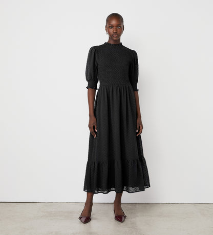 A woman stands against a white wall wearing the Amelia Black Chiffon Dress, featuring puff sleeves and a ruffled hem. She completes her look with pointed dark heels and exudes a confident, composed expression.