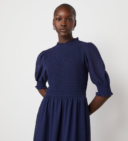 A woman wearing the Amelia Navy Georgette Midi Dress, featuring a high neckline, subtle dot pattern, and shirred bodice, stands confidently against a plain white background.