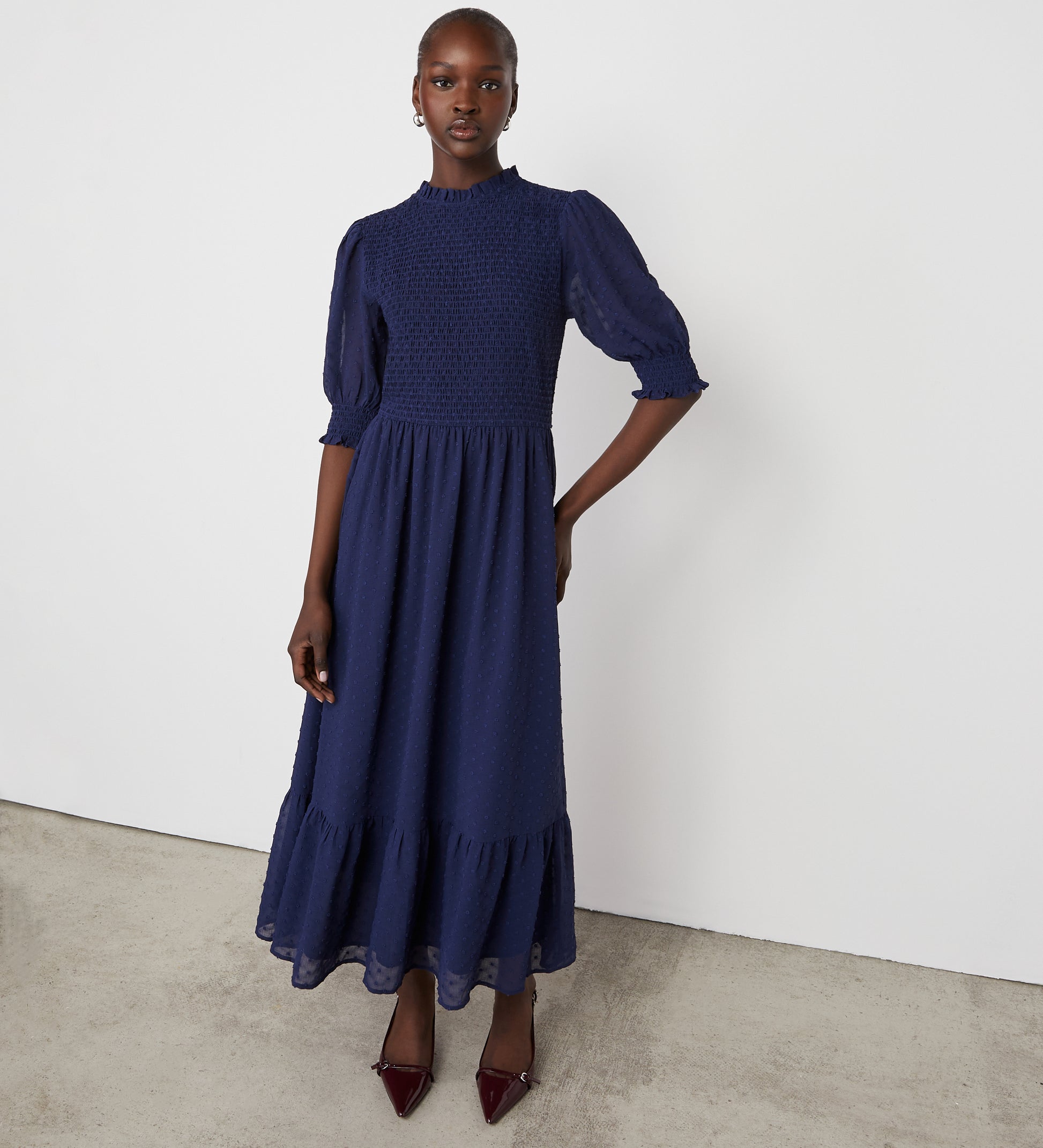 A woman stands against a white wall, wearing the Amelia Navy Georgette Midi Dress featuring puffed half sleeves and a ruffled hem, paired with dark pointed-toe shoes.