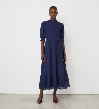 A woman stands against a white wall wearing the Amelia Navy Georgette Midi Dress with puffed half sleeves, a shirred bodice, ruffle hem detail, and burgundy heeled shoes.