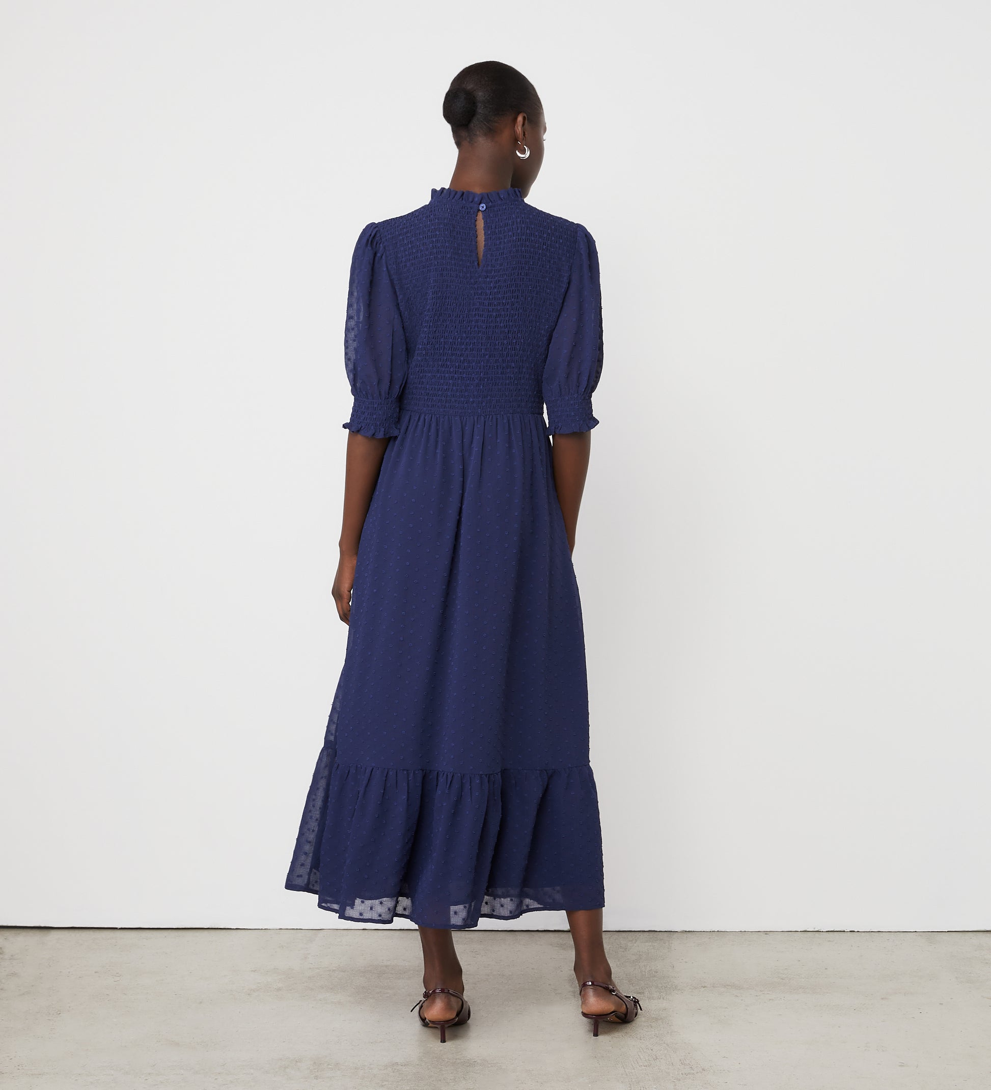 A woman stands facing away on a plain background, wearing the Amelia Navy Georgette Midi Dress with long sleeves, high neck, and ruffled hem.