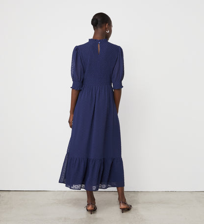 A woman stands facing away on a plain background, wearing the Amelia Navy Georgette Midi Dress with long sleeves, high neck, and ruffled hem.