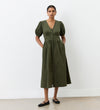 A woman stands against a white wall wearing the Arlow Khaki Corduroy Midi Dress, featuring short sleeves, button-through front, V-neckline, and pockets—ideal for autumn. She has her hands in the pockets and pairs it with classic black flats.