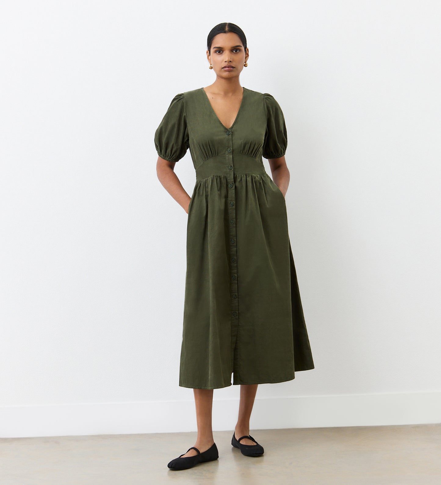 A woman stands against a white wall wearing the Arlow Khaki Corduroy Midi Dress, featuring short sleeves, button-through front, V-neckline, and pockets—ideal for autumn. She has her hands in the pockets and pairs it with classic black flats.