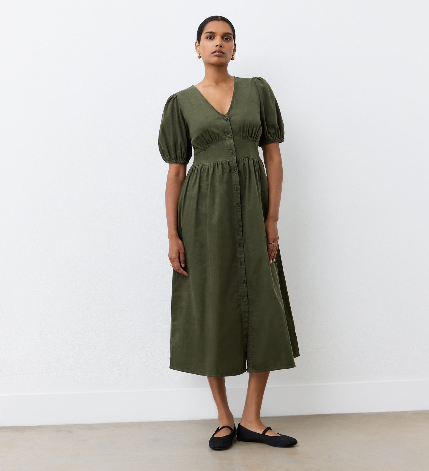 A woman stands against a white wall, wearing the Arlow Khaki Corduroy Midi Dress with short puff sleeves and black flats. Her hair is pulled back and she looks straight ahead, showcasing effortless autumn style.