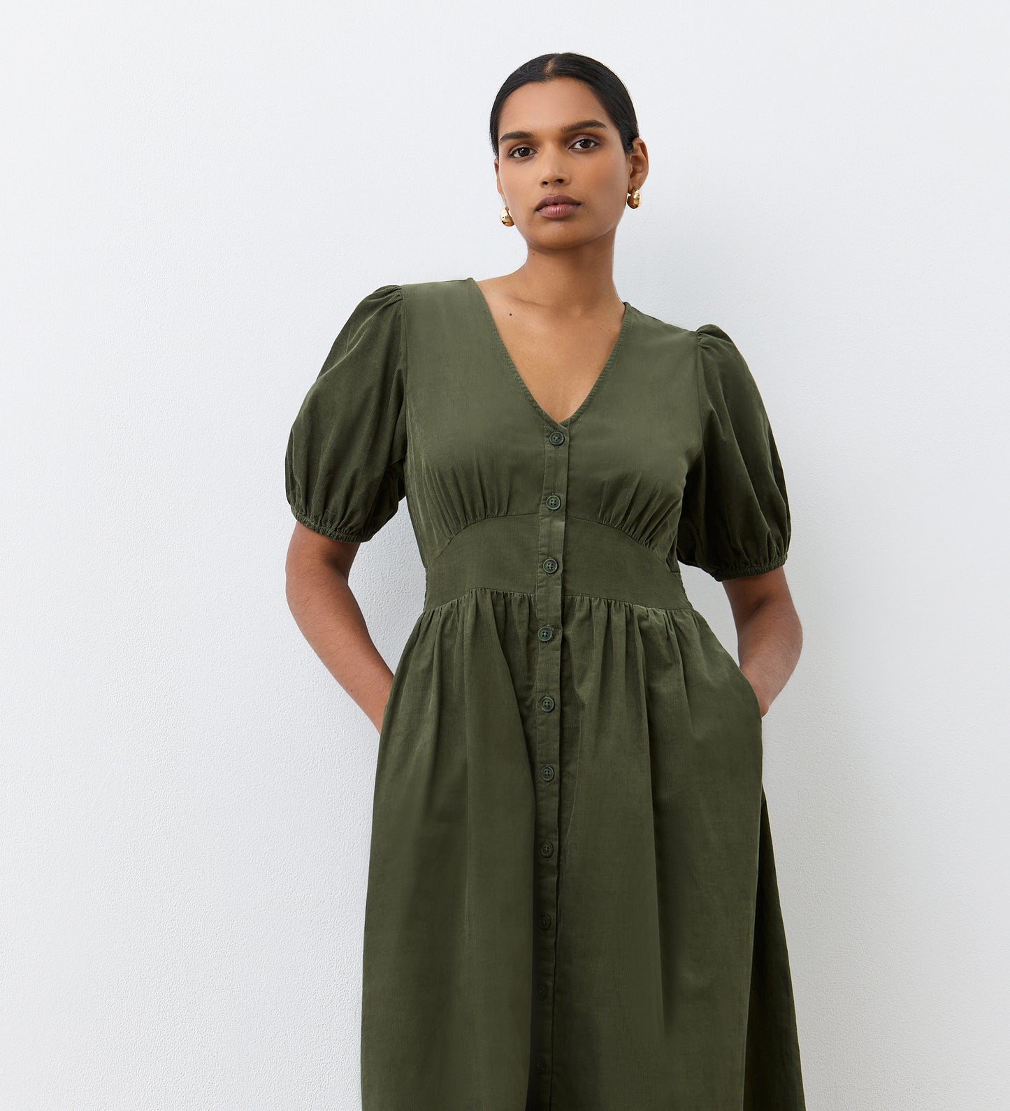 A woman stands against a white background in the Arlow Khaki Corduroy Midi Dress, featuring a V-neck, button-through front, puffed sleeves, and pockets—an effortless autumn option. Her short hair is pulled back.