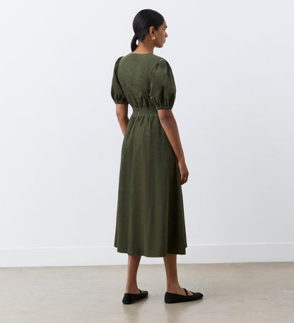 A woman stands indoors facing away, wearing the Arlow Khaki Corduroy Midi Dress with short sleeves and an elastic waist, paired with black flats—an effortless autumn look against a plain white wall and light floor.