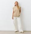 A woman with long blonde hair stands against a white wall wearing the Ava Beige Sweater Vest, wide-leg white pants, and beige shoes. With a relaxed pose and neutral expression, her chic layered look is perfect for any occasion.