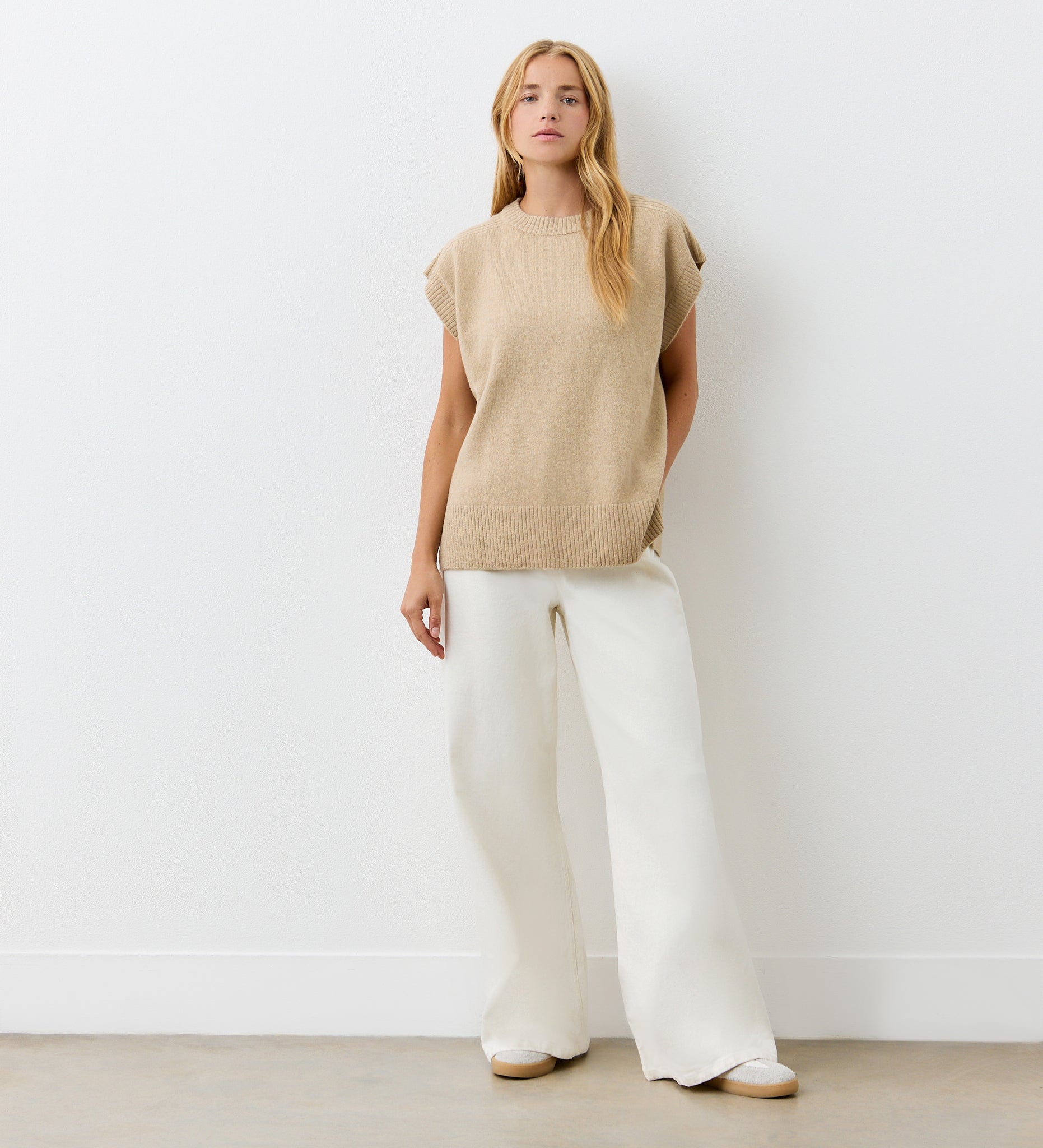 A woman with long blonde hair stands against a white wall wearing the Ava Beige Sweater Vest, wide-leg white pants, and beige shoes. With a relaxed pose and neutral expression, her chic layered look is perfect for any occasion.