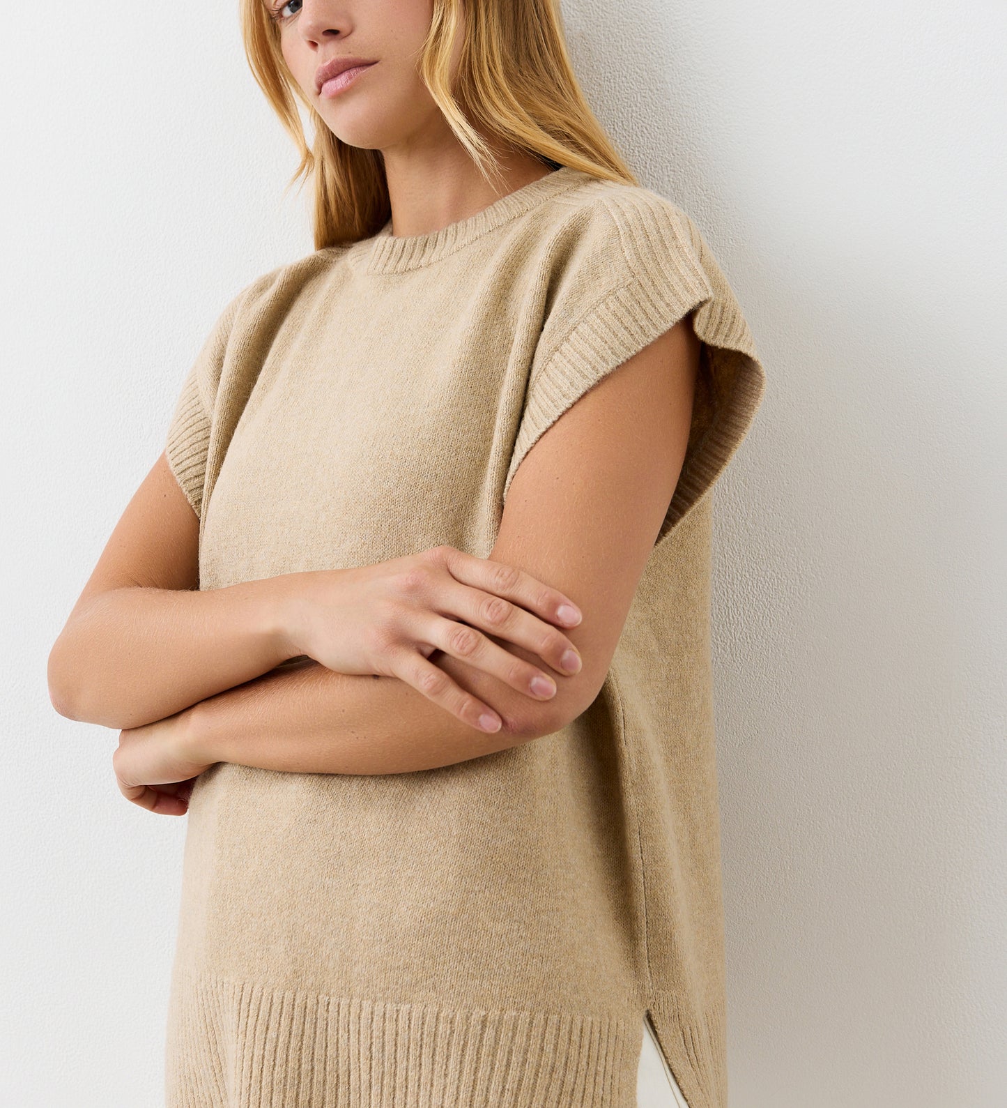A woman with long blonde hair stands against a white wall, wearing the Ava Beige Sweater Vest. Her arms are crossed loosely, she has a neutral expression, and only her upper body is visible.