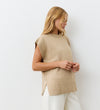 A woman with long blonde hair wears the sleeveless Ava Beige Sweater Vest and white pants, standing against a plain white background while looking down—a chic, effortless look for layering.