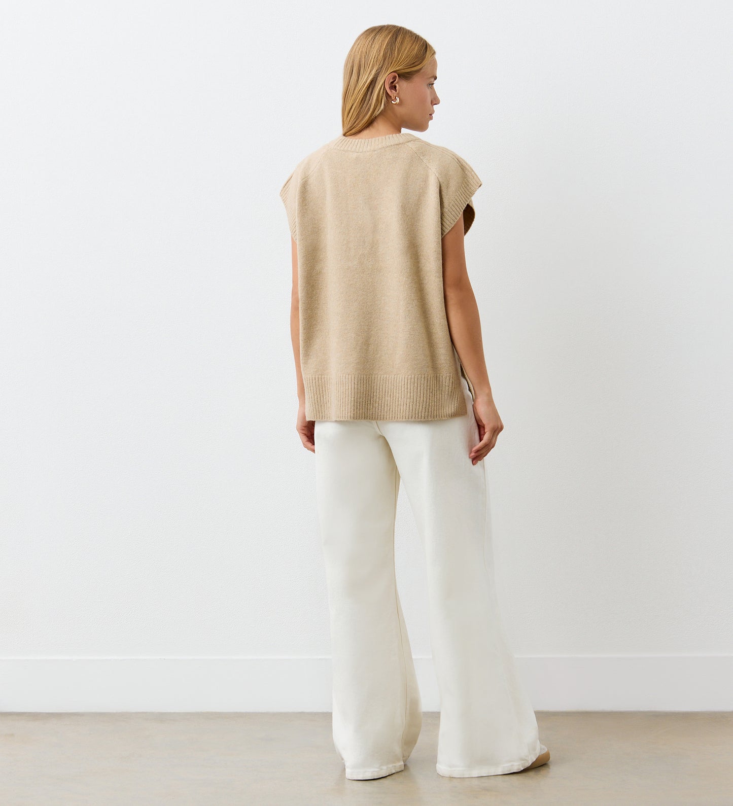 A woman stands with her back to the camera, wearing the Ava Beige Sweater Vest over wide-legged white pants, set against a plain white wall and concrete floor.