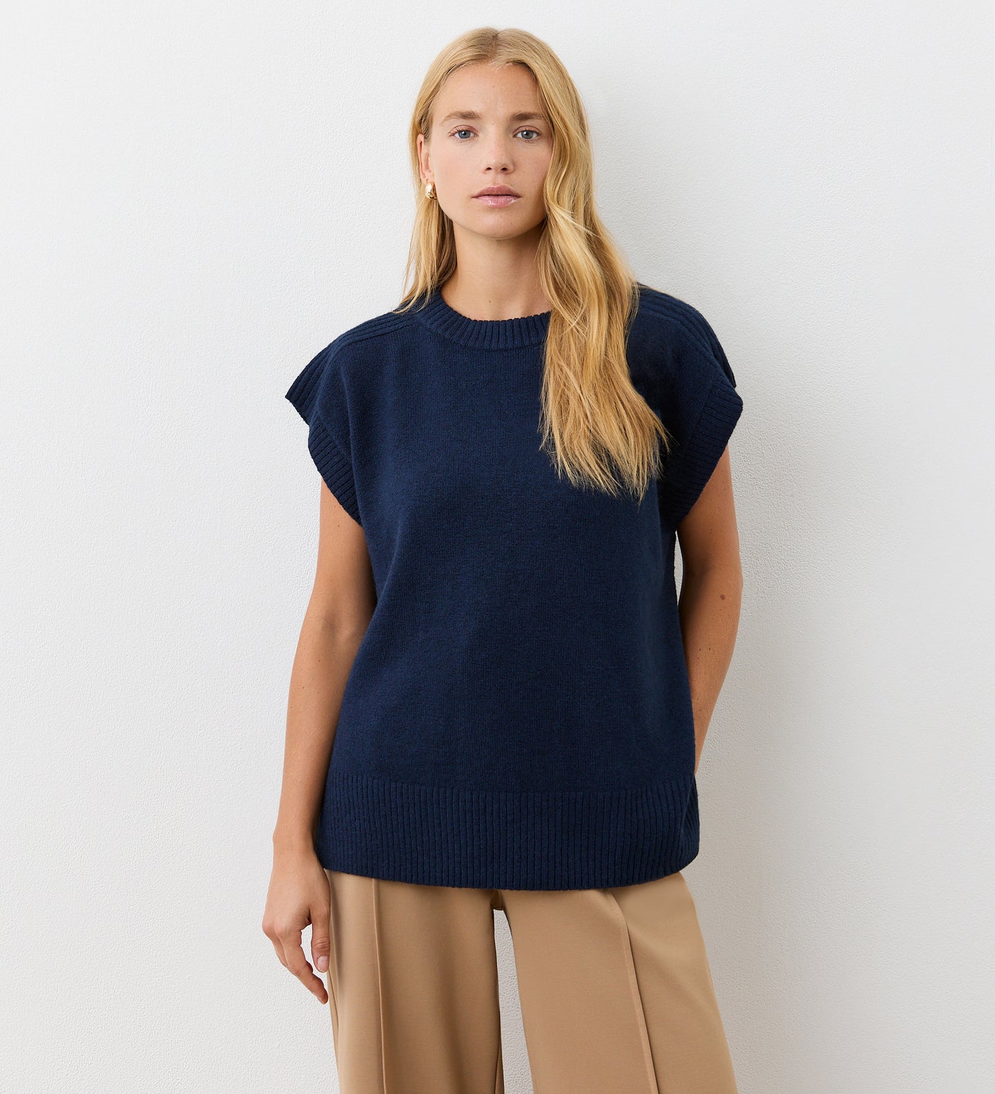 A woman with long blonde hair wears the Ava Navy Knitted Vest with beige trousers, standing against a plain white background and looking calmly at the camera—an effortless look for autumn.