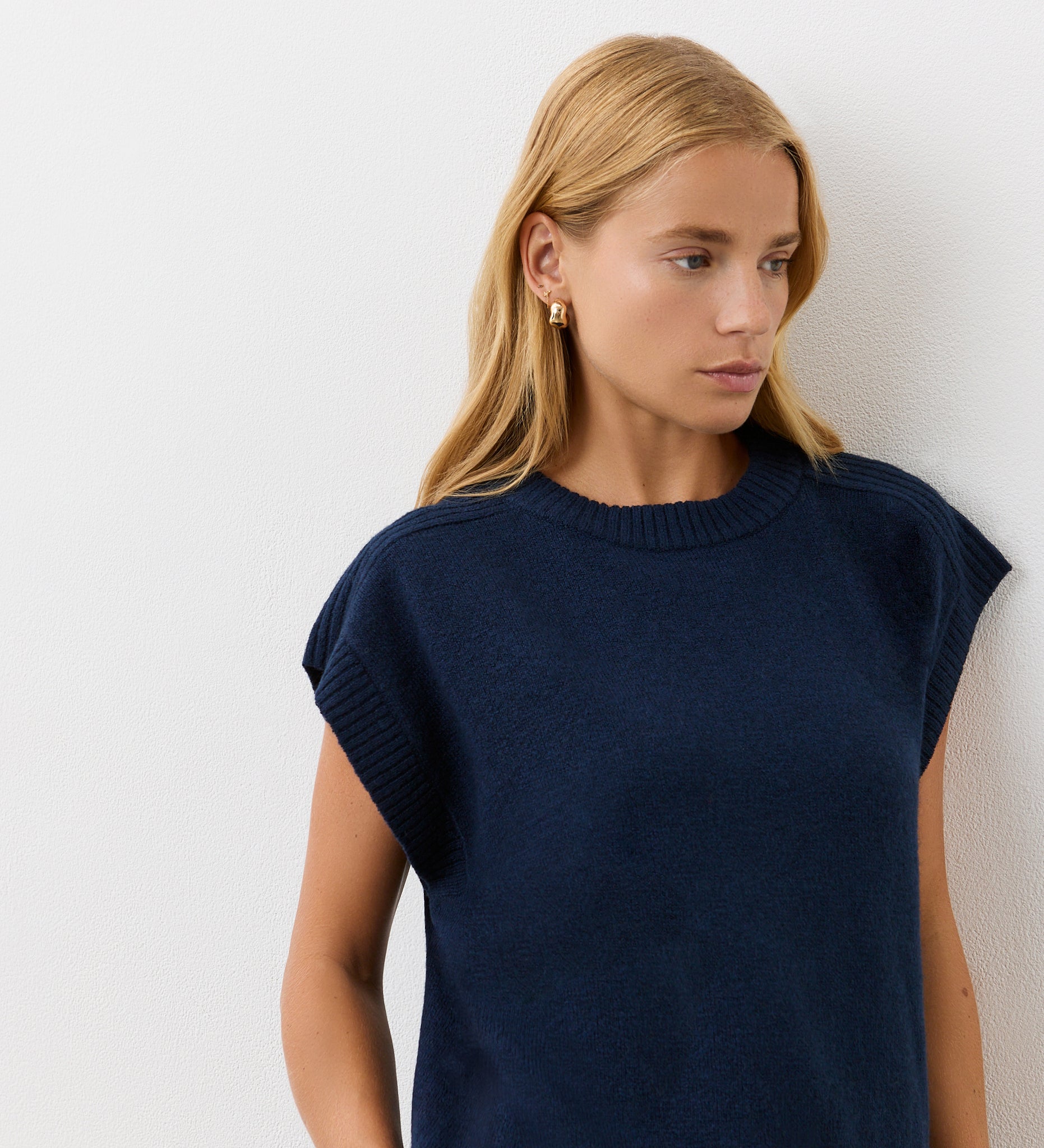 A woman with long blonde hair wears the Ava Navy Knitted Vest—a short-sleeved design—while standing against a white wall and looking slightly left, showcasing this effortless layer for autumn.