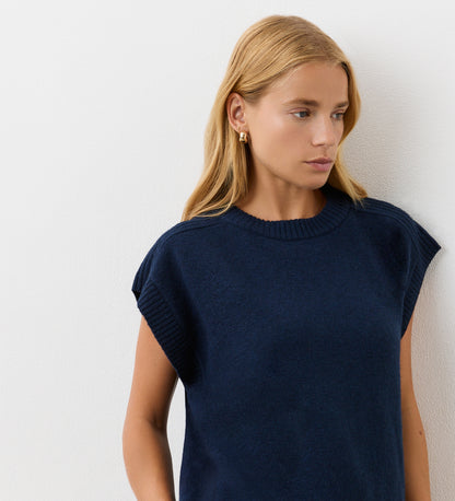A woman with long blonde hair wears the Ava Navy Knitted Vest—a short-sleeved design—while standing against a white wall and looking slightly left, showcasing this effortless layer for autumn.