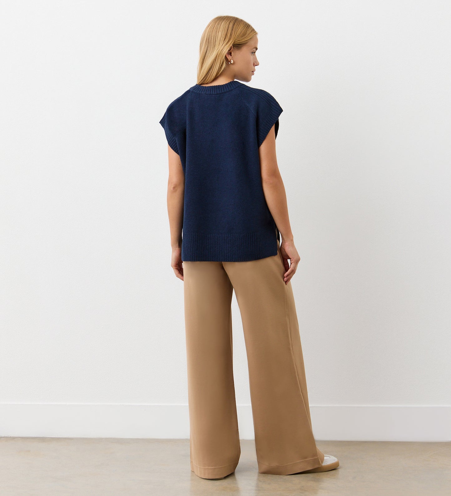 A woman with straight blonde hair stands facing away from the camera, indoors against a plain white wall, wearing the Ava Navy Knitted Vest and wide-leg tan pants for an effortless autumn layered look.