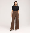 A woman with long, straight hair and a neutral expression stands against a plain background wearing a black short-sleeve top, Addie Mushroom Gaberdine Twill Wide Leg Trousers in brown, and black pointed-toe shoes.