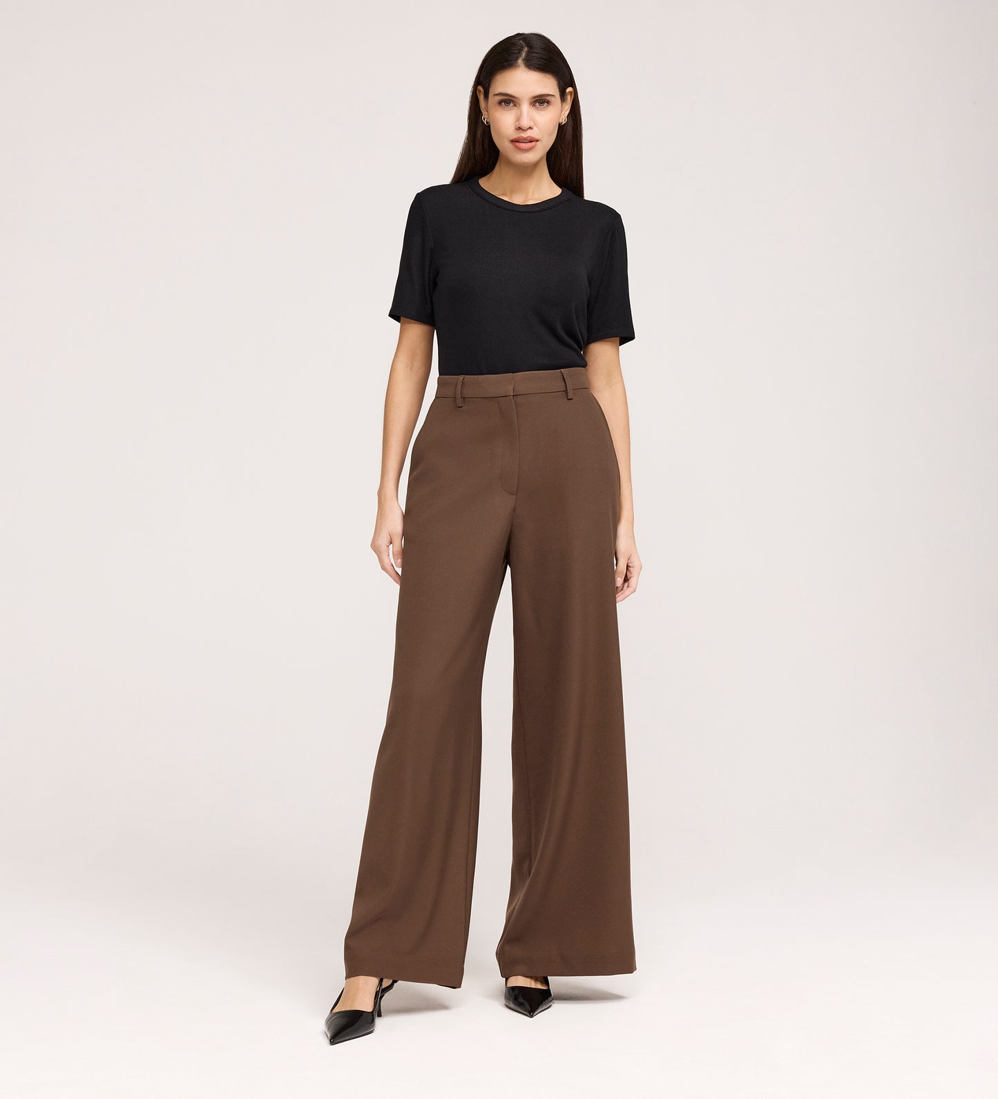 A woman with long, straight hair and a neutral expression stands against a plain background wearing a black short-sleeve top, Addie Mushroom Gaberdine Twill Wide Leg Trousers in brown, and black pointed-toe shoes.
