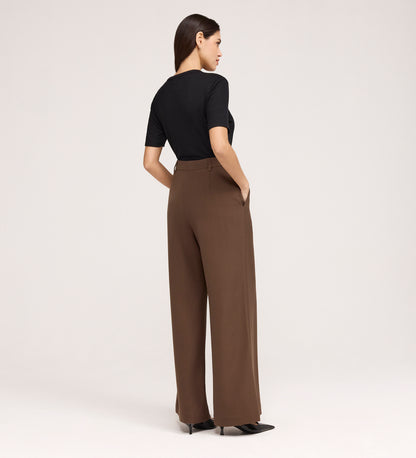 A woman stands with her back to the camera, wearing a black short-sleeve top and the Addie Mushroom Gaberdine Twill Wide Leg Trousers, hands in pockets, against a plain light background.