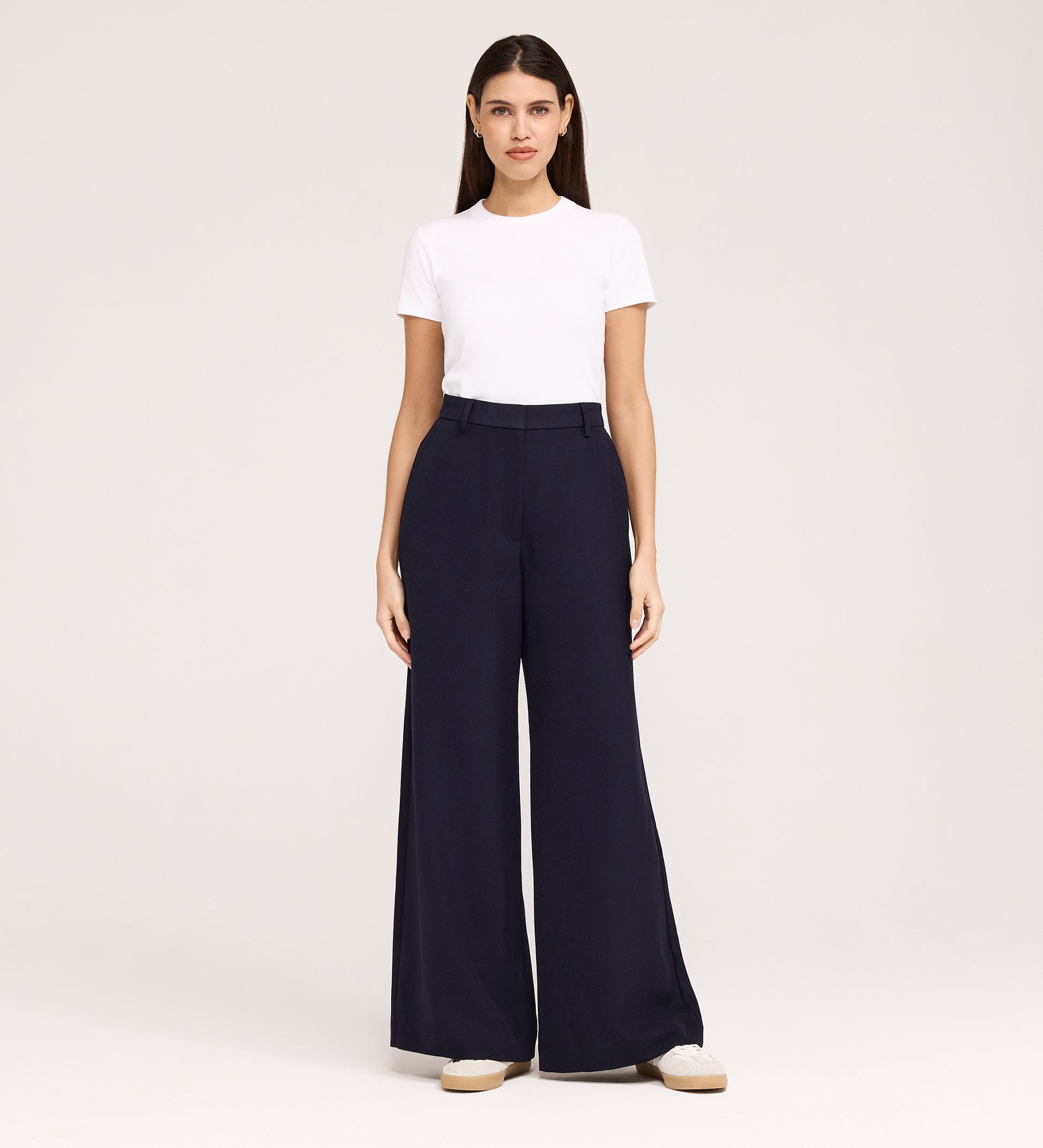 A woman faces forward against a plain light background, wearing a white short-sleeve t-shirt, the Addie Navy Gaberdine Twill Wide Leg Trousers, and white shoes.