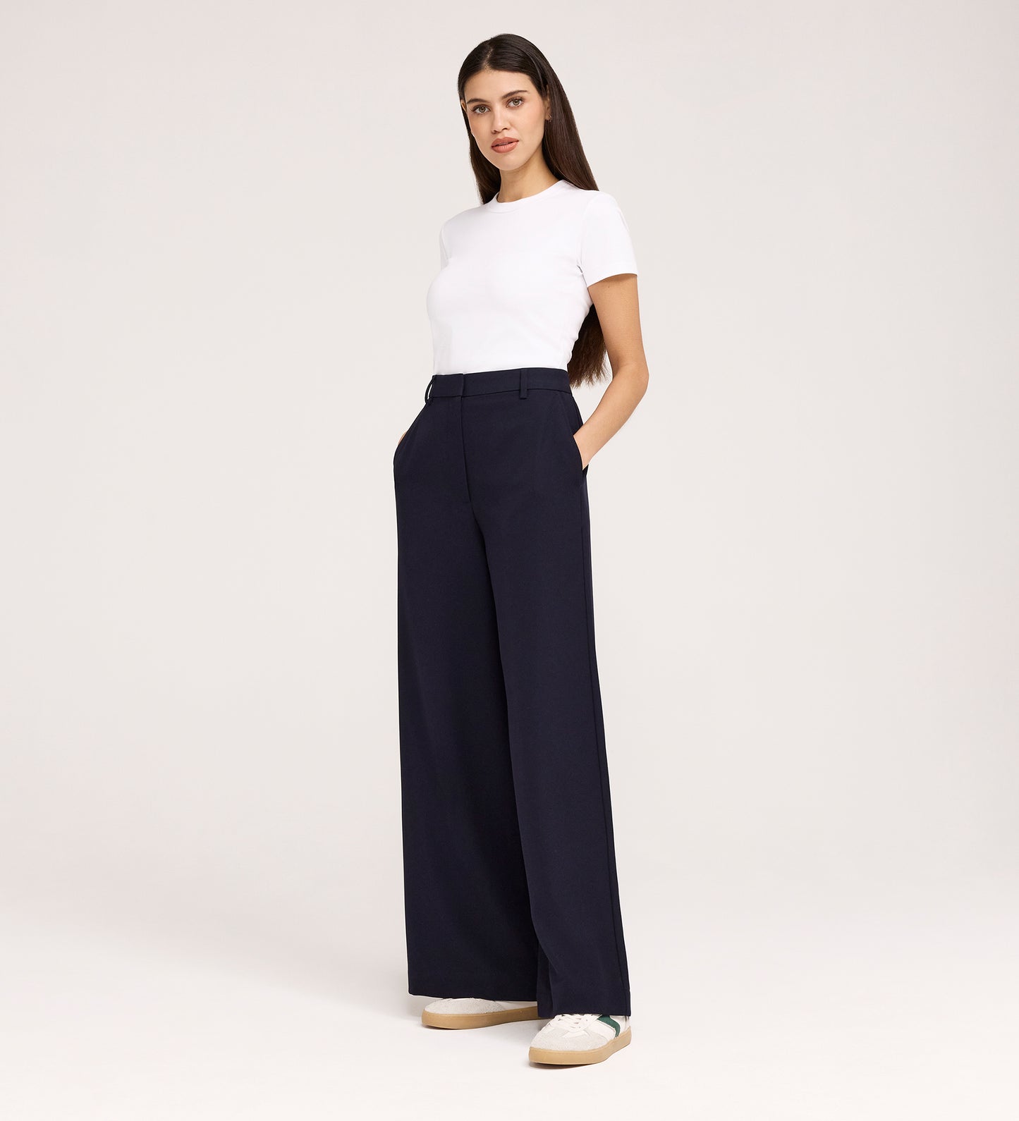 A woman with long brown hair stands in a studio wearing a fitted white t-shirt, the Addie Navy Gaberdine Twill Wide Leg Trousers, and white sneakers. She poses with her hands in her pockets against a plain background.