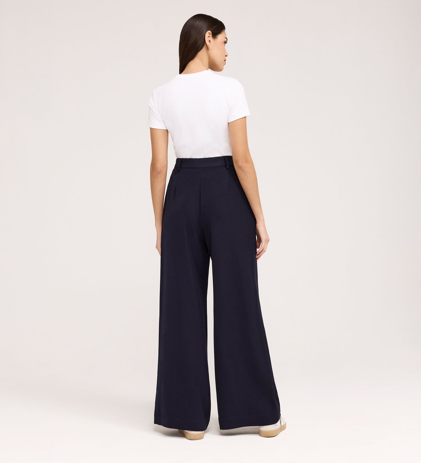 A woman stands facing away in a white short-sleeve t-shirt tucked into Addie Navy Gaberdine Twill Wide Leg Trousers with beige shoes, against a plain light background.