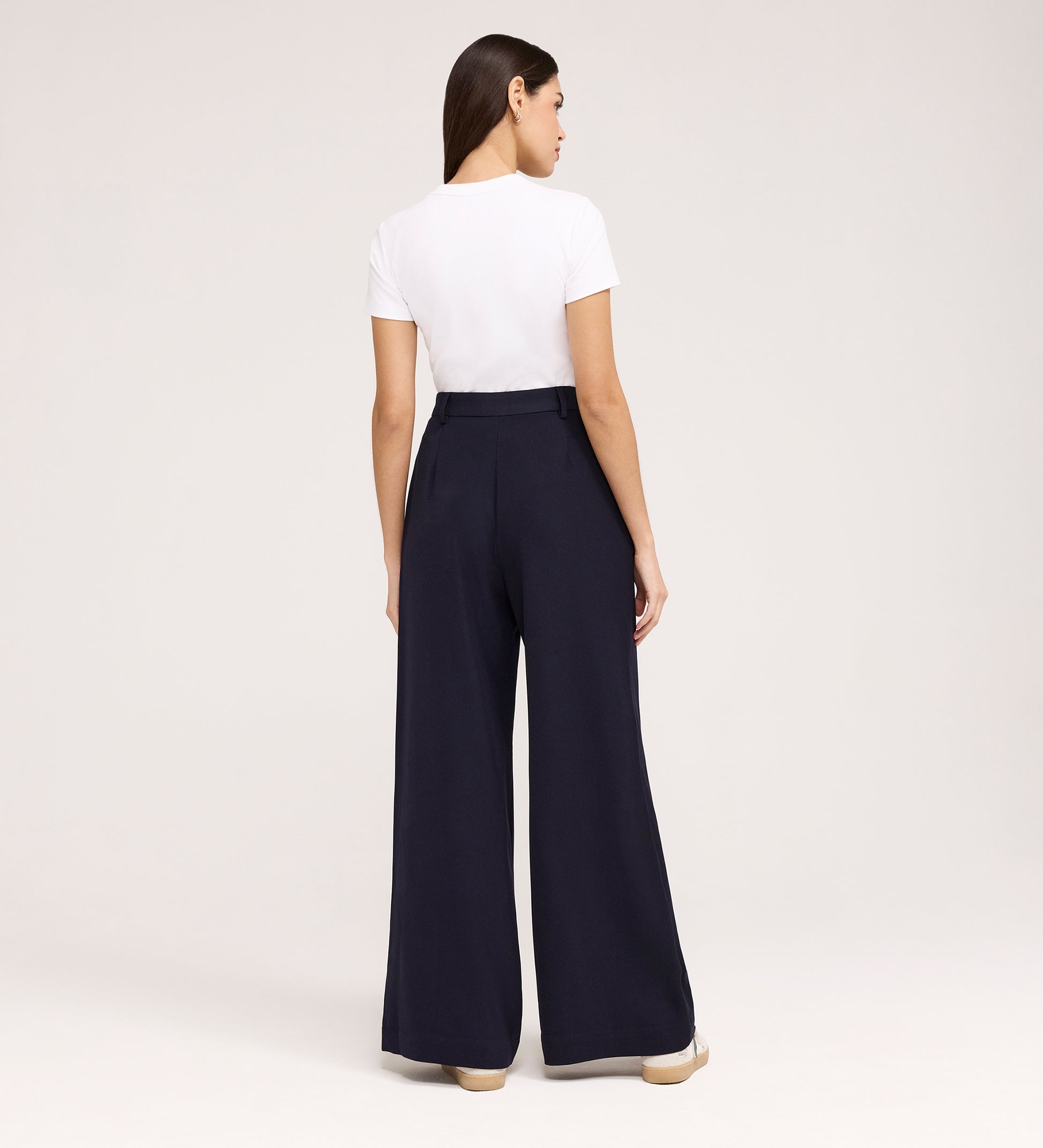 A woman stands facing away in a white short-sleeve t-shirt tucked into Addie Navy Gaberdine Twill Wide Leg Trousers with beige shoes, against a plain light background.