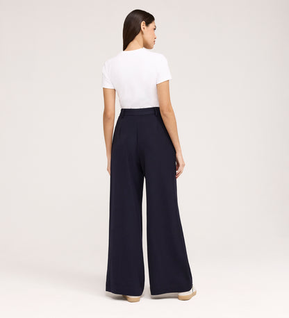 A woman stands facing away in a white short-sleeve t-shirt tucked into Addie Navy Gaberdine Twill Wide Leg Trousers with beige shoes, against a plain light background.