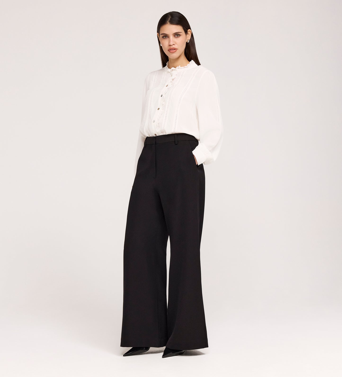 A woman stands against a plain background wearing a white ruffled-collar blouse and Addie Black Gaberdine Twill Wide Leg Trousers, paired with black pointed-toe shoes.