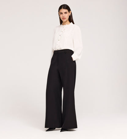 A woman stands against a plain background wearing a white ruffled-collar blouse and Addie Black Gaberdine Twill Wide Leg Trousers, paired with black pointed-toe shoes.