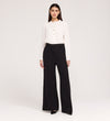 A woman stands against a plain white background wearing a white long-sleeve blouse with buttons, Addie Black Gaberdine Twill Wide Leg Trousers, and black heels.