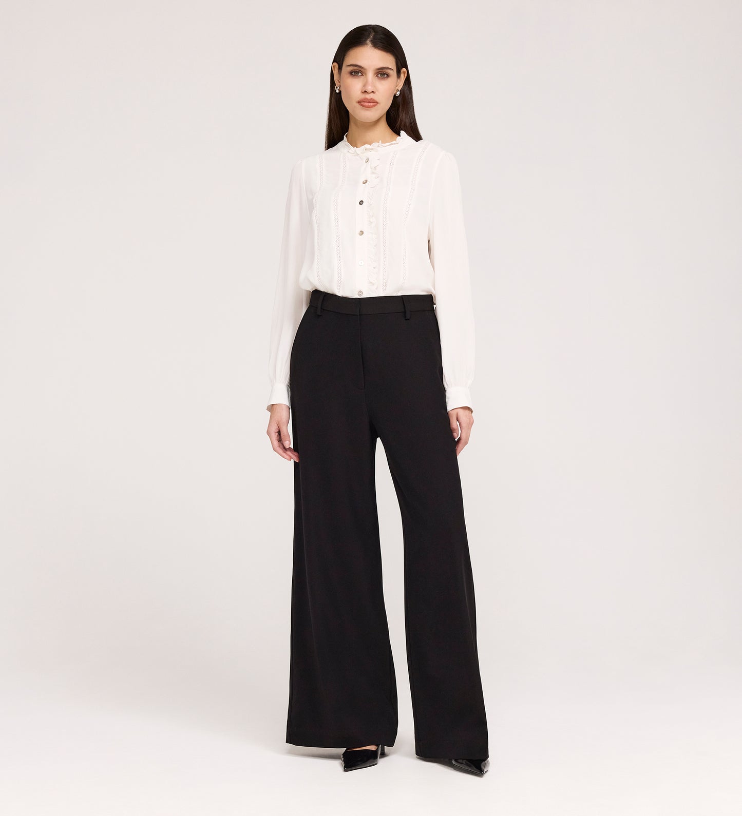 A woman stands against a plain white background wearing a white long-sleeve blouse with buttons, Addie Black Gaberdine Twill Wide Leg Trousers, and black heels.