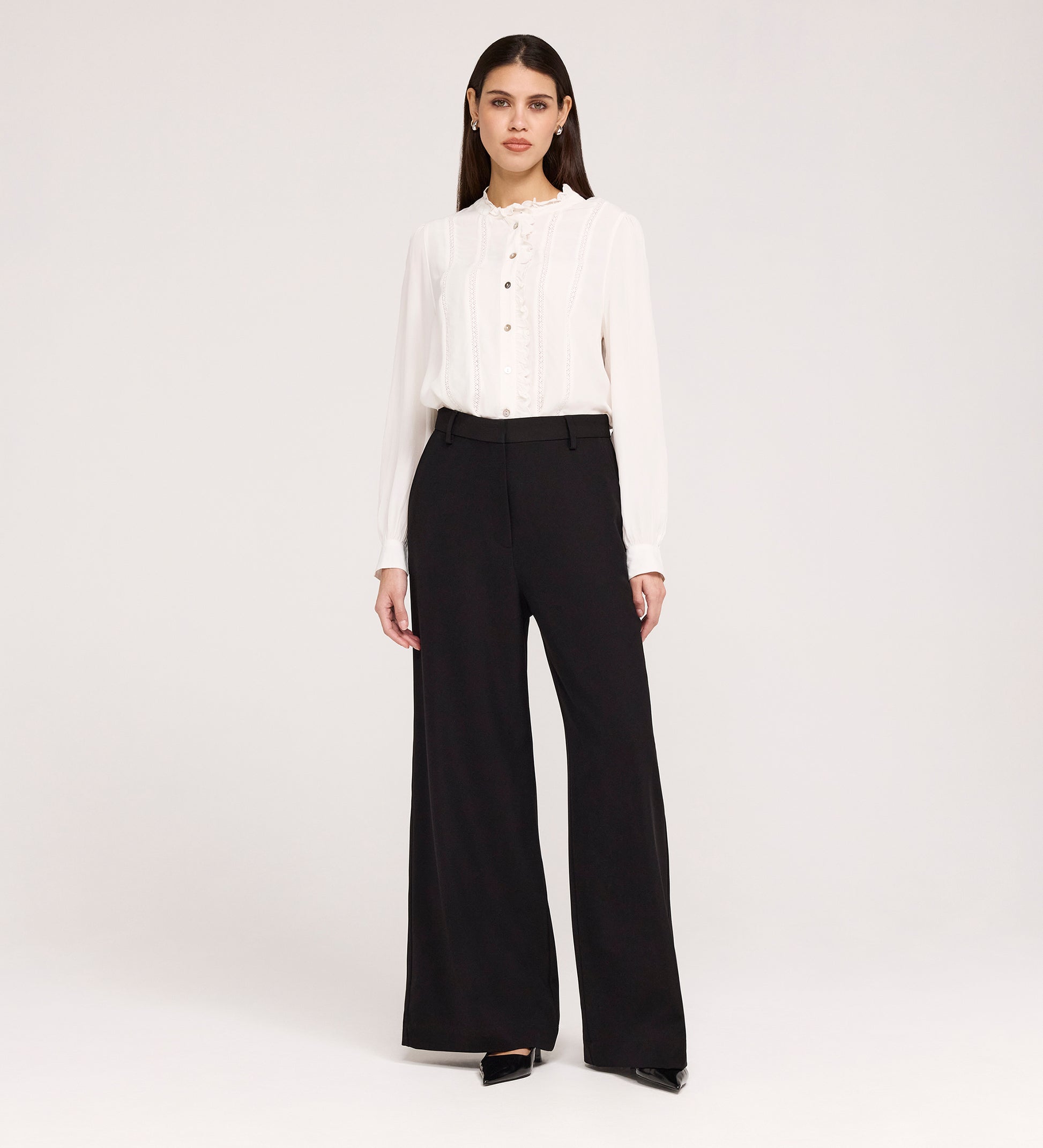 A woman stands against a plain white background wearing a white long-sleeve blouse with buttons, Addie Black Gaberdine Twill Wide Leg Trousers, and black heels.