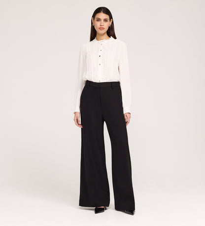 A woman stands against a plain white background wearing a white long-sleeve blouse with buttons, Addie Black Gaberdine Twill Wide Leg Trousers, and black heels.