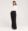 A woman stands against a plain background, wearing high-waisted Addie Black Gaberdine Twill Wide Leg Trousers, a white ruffle long-sleeve blouse, and black pointed-toe shoes with her hands in her pockets.