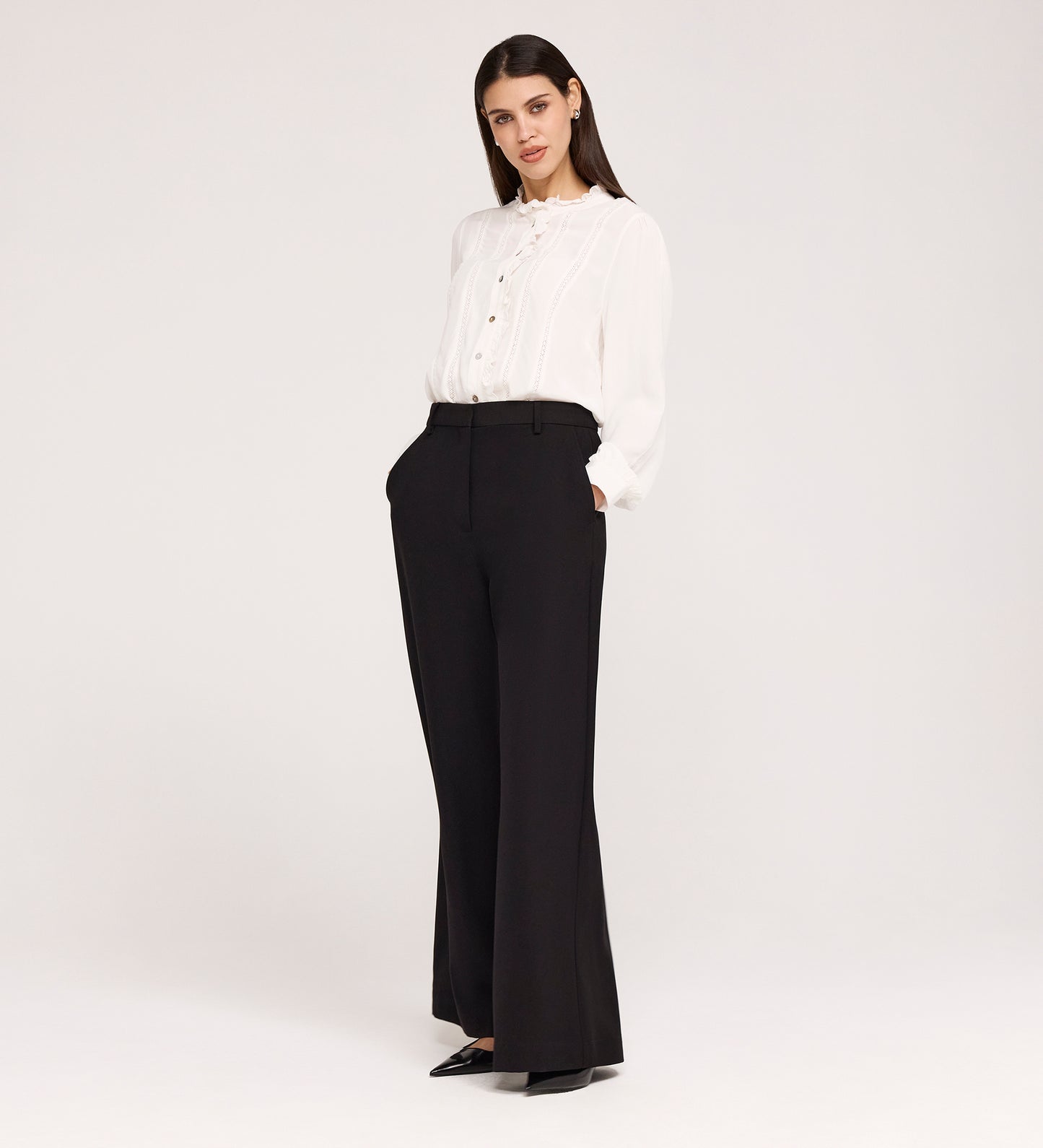 A woman stands against a plain background, wearing high-waisted Addie Black Gaberdine Twill Wide Leg Trousers, a white ruffle long-sleeve blouse, and black pointed-toe shoes with her hands in her pockets.
